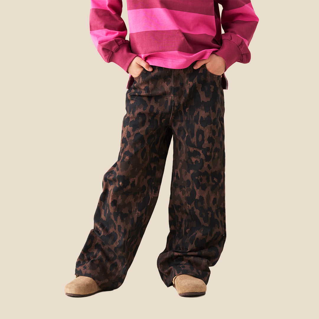 A person wearing loose, wide-leg brown and black leopard-print pants, paired with a bright pink striped long-sleeve top and tan slip-on shoes, against a plain beige background.