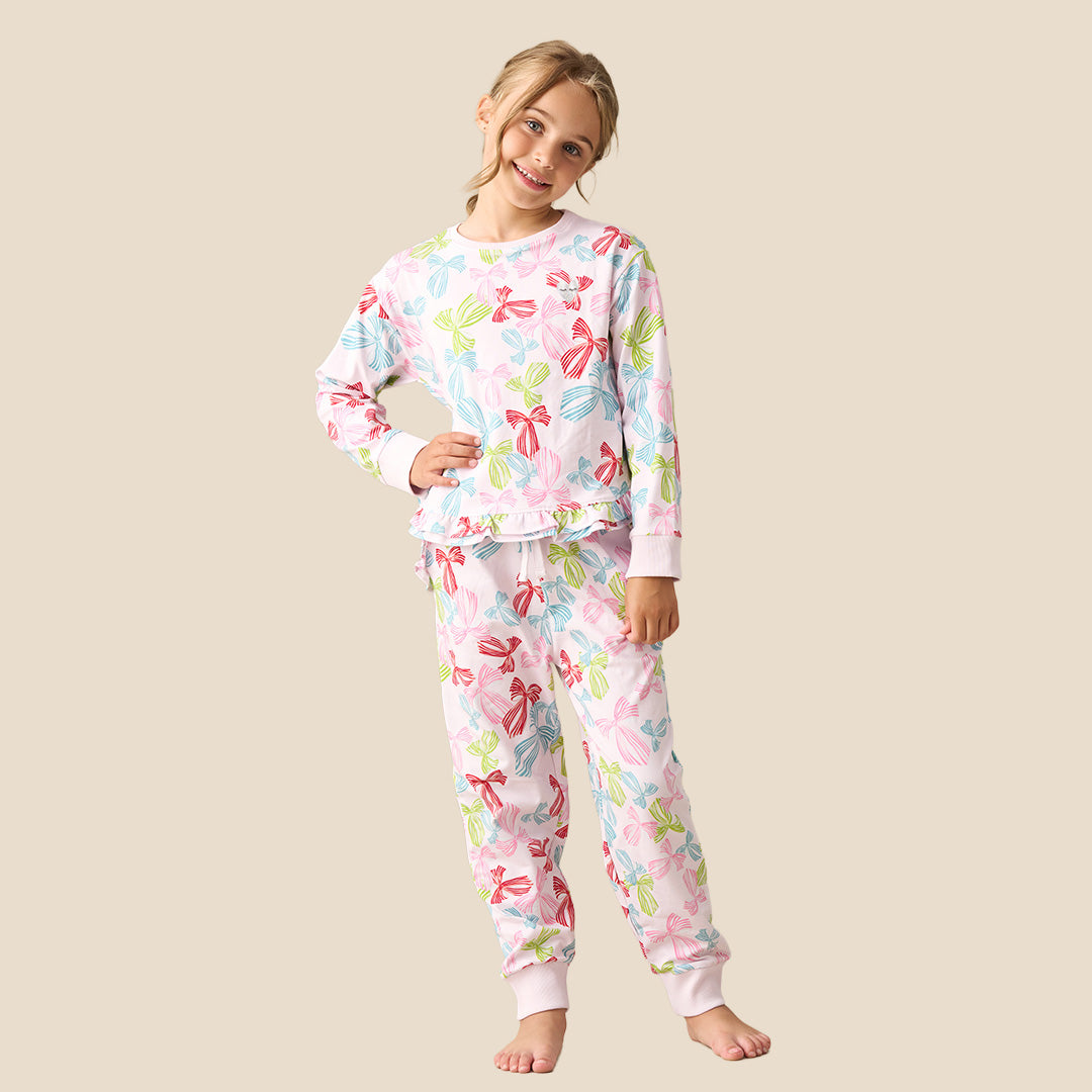 A young girl modeling a two-piece pajama set featuring a long-sleeve top and matching pants with a colorful bow pattern on a light pink background, set against a solid beige backdrop.