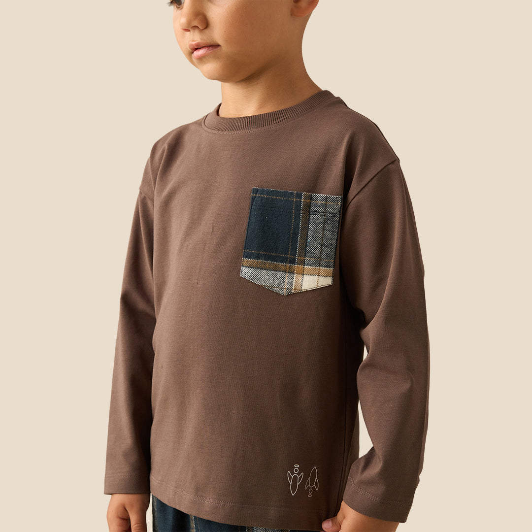 A young boy modeling a two-piece pajama set consisting of a brown long-sleeve shirt with a plaid chest pocket and matching plaid pants, set against a solid beige background.