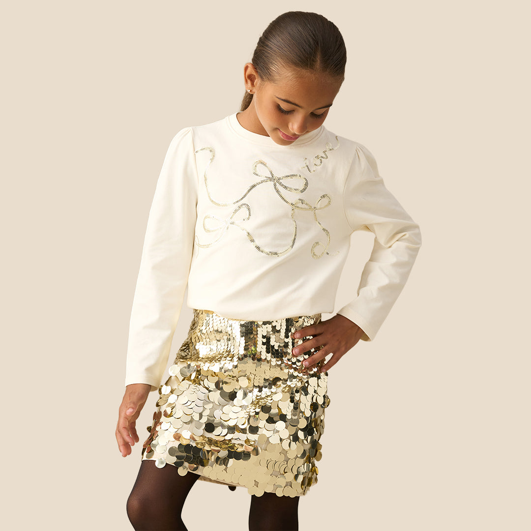 A close-up of a person wearing a shiny gold sequin mini skirt with a matching gold chain belt, paired with a cream-colored top and black tights, against a solid beige background.