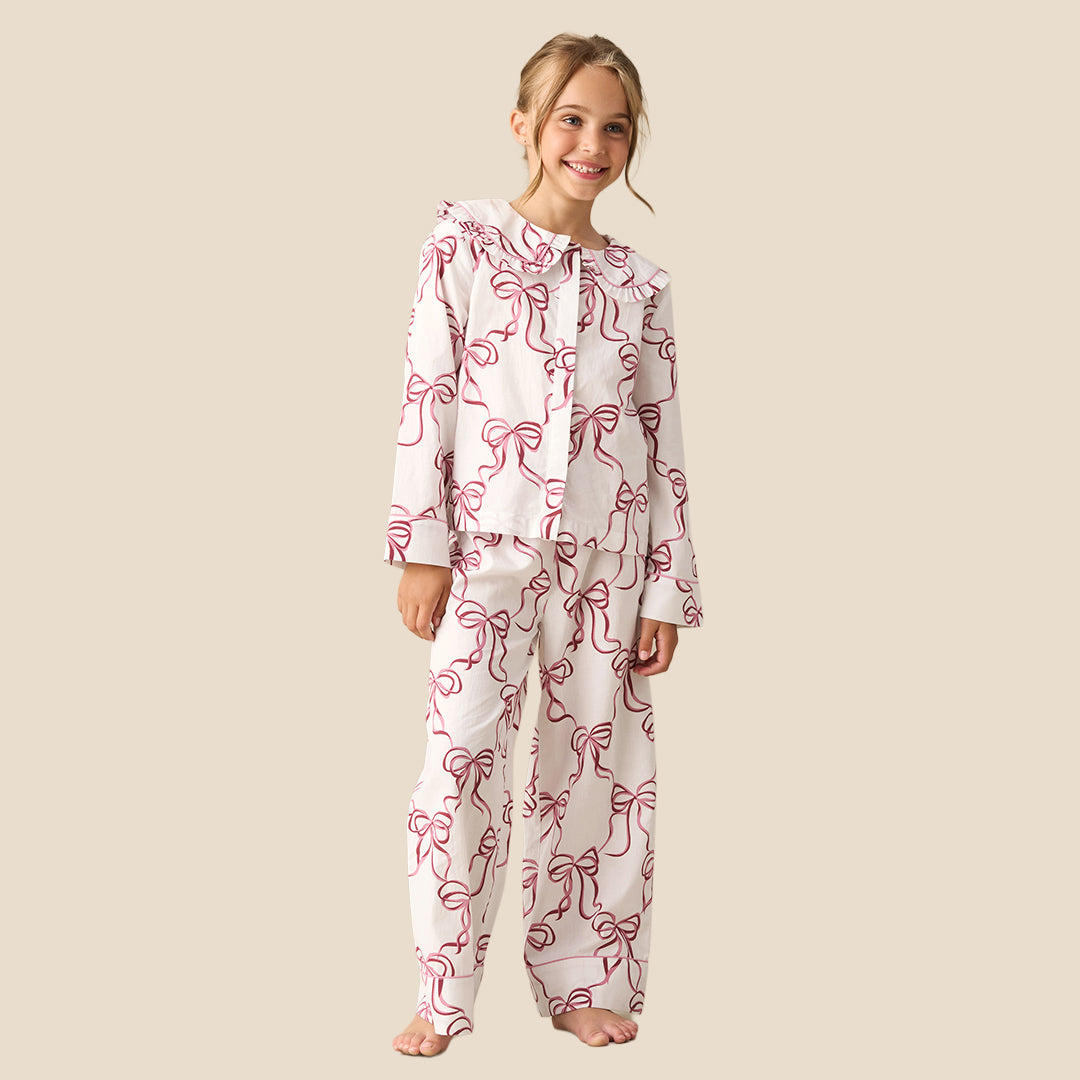 A young girl modeling a two-piece pajama set with a white base and a pink bow pattern, standing against a solid beige background.