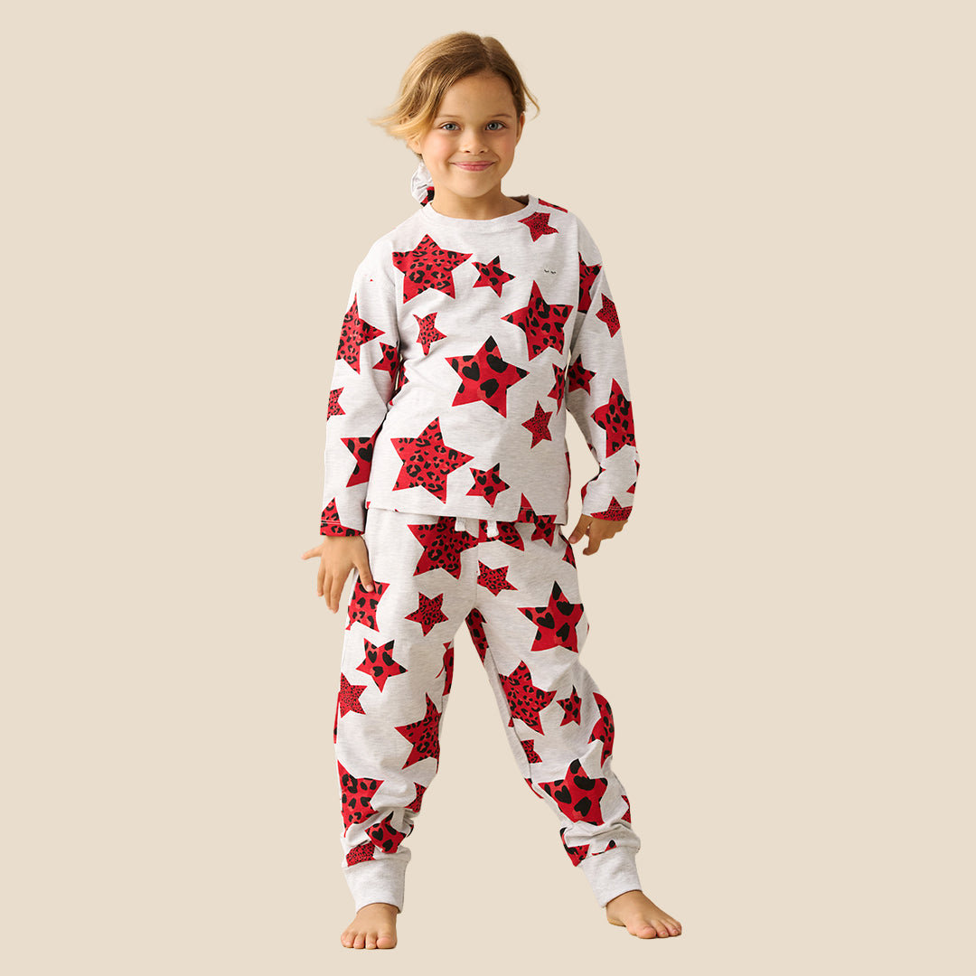 A young girl modeling a matching two-piece pajama set with a white base and a red star and ladybug print, set against a solid beige background.