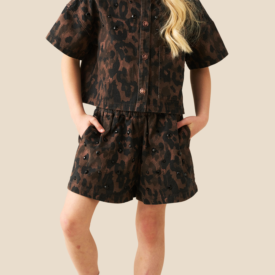 A child wearing a matching short-sleeve button-front top and shorts set in a dark brown and black leopard print, with decorative pearl-like embellishments, photographed against a plain beige background.