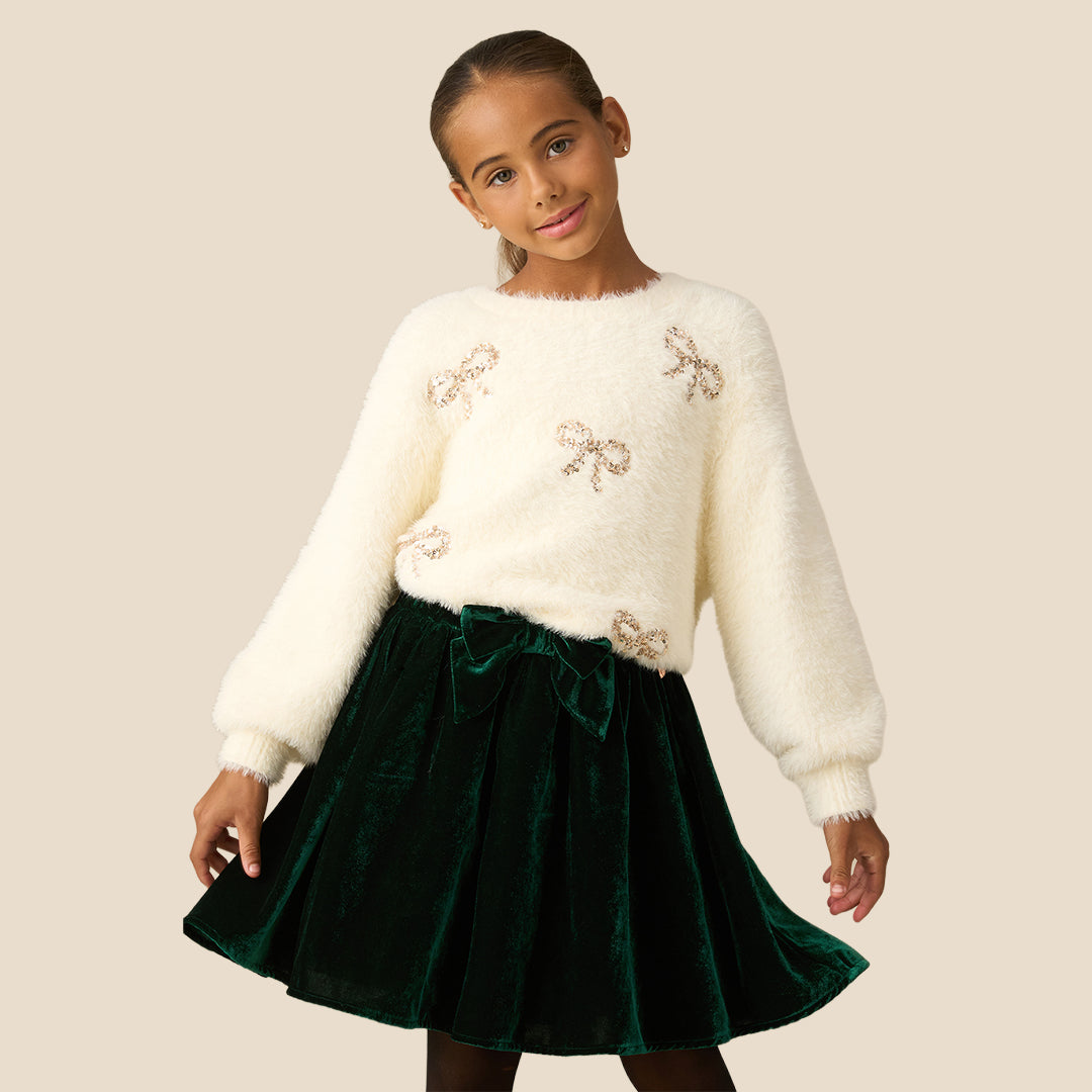 A young girl with her hair in a ponytail wearing a fuzzy off-white long-sleeve sweater with sequined bow details and a dark green velvet skirt, set against a solid beige background.