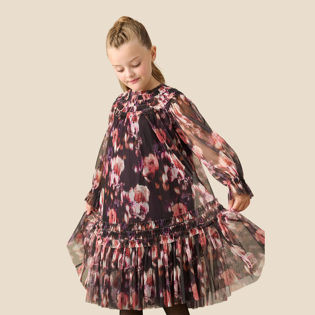 A young girl with her hair in a ponytail is wearing a long-sleeved, tiered tulle dress with a pink, red, and white floral pattern on a dark background, set against a solid beige studio backdrop.