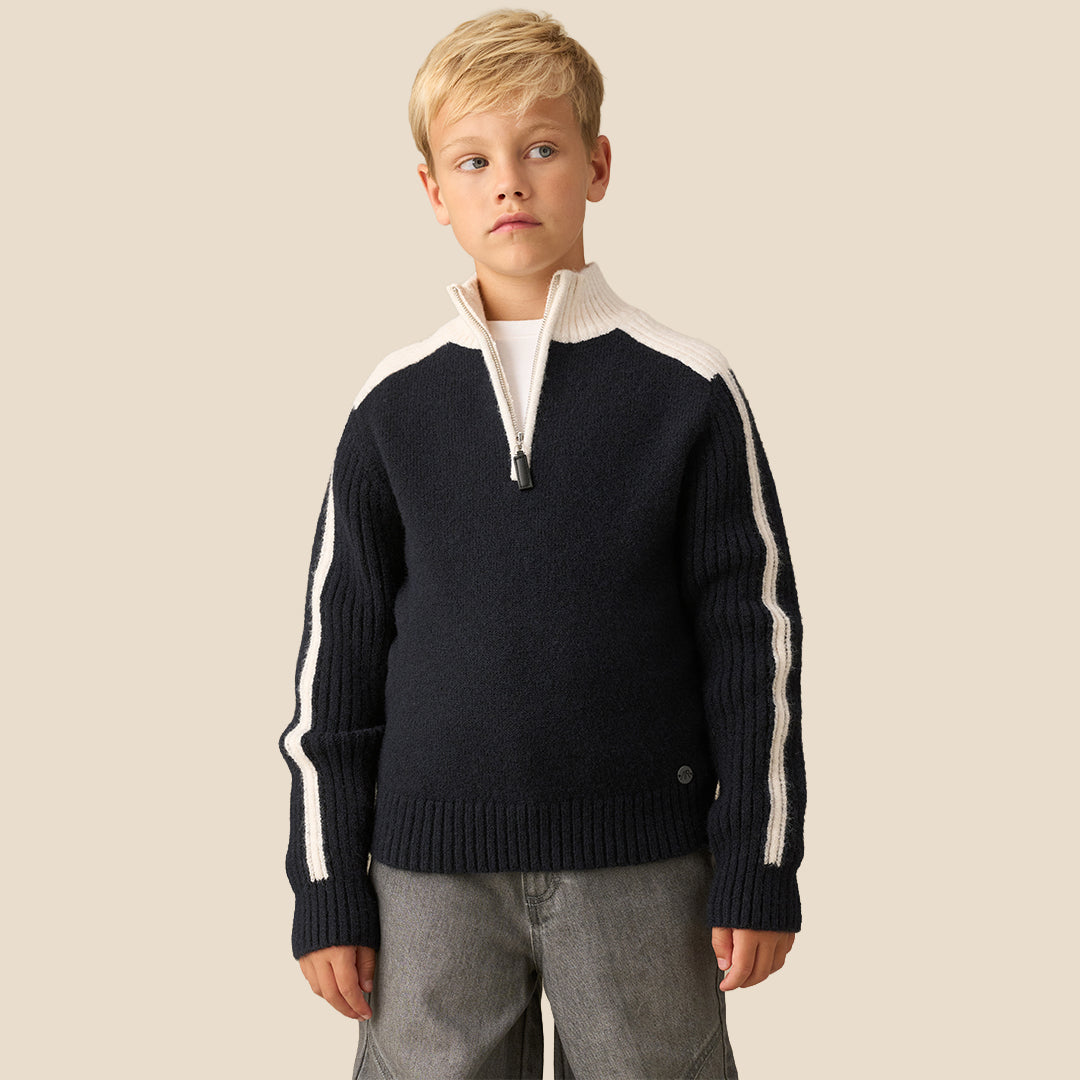 A young boy wearing a navy blue and cream knit quarter-zip sweater with white stripe details on the sleeves, paired with gray pants, against a solid beige background.