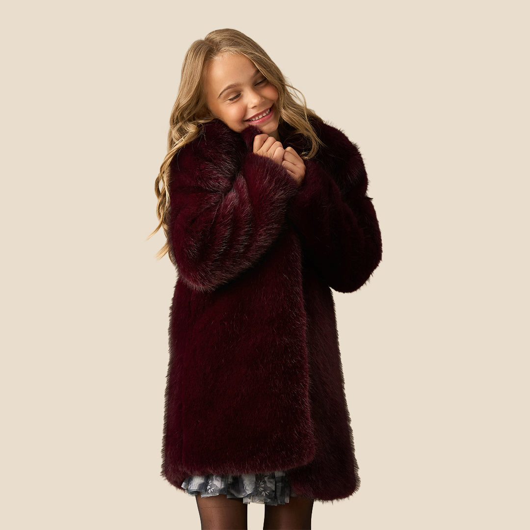 A young girl with blonde hair wearing a plush, dark burgundy faux fur coat over a dark patterned dress, standing against a solid beige background.