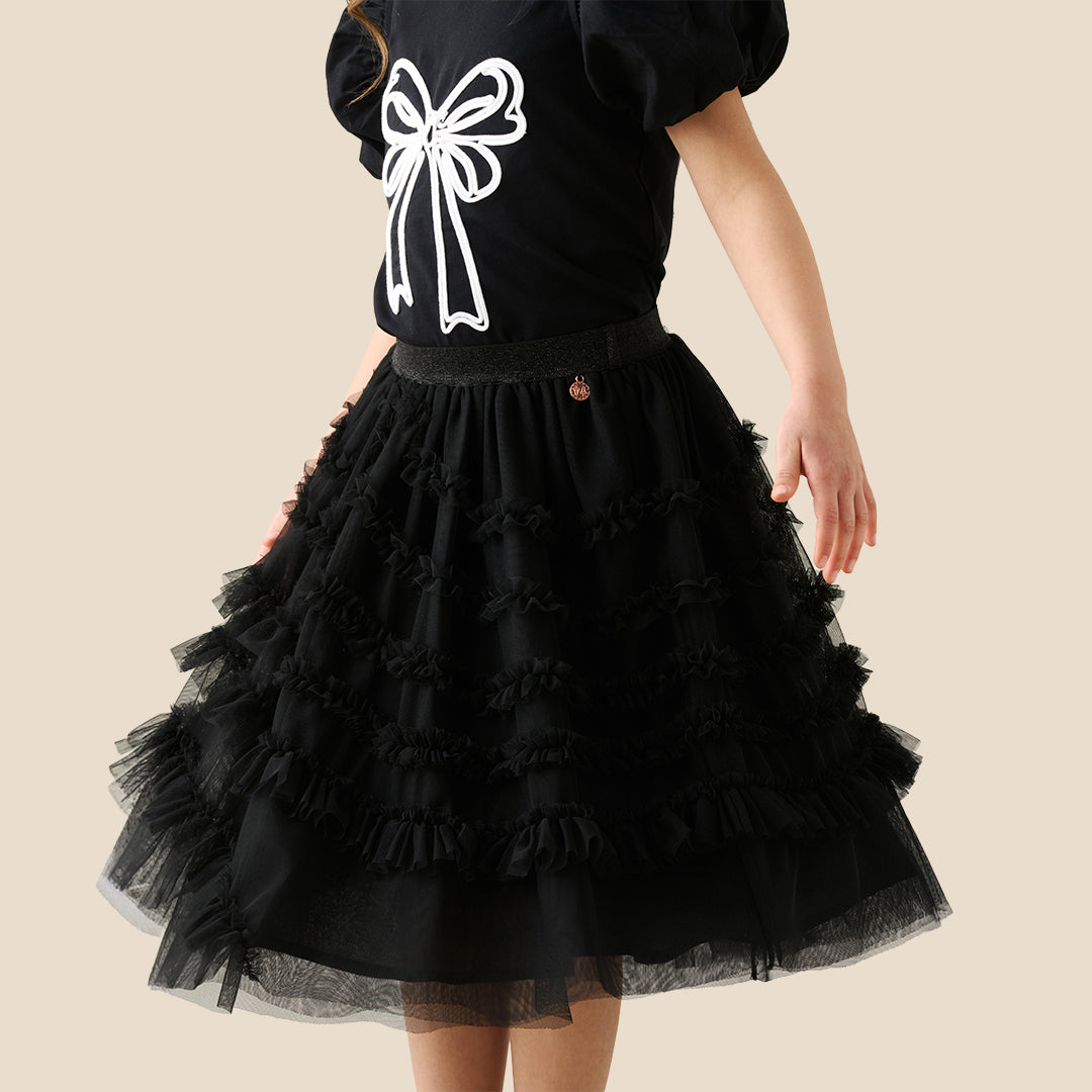 A child wearing a black puff-sleeve top with a large white bow graphic and a layered black ruffled tulle skirt, shown against a plain beige studio background.