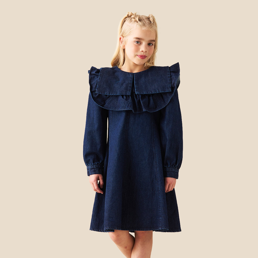 A young girl with blonde hair in two buns wearing a dark blue denim dress with a large ruffled collar, standing against a solid beige background.