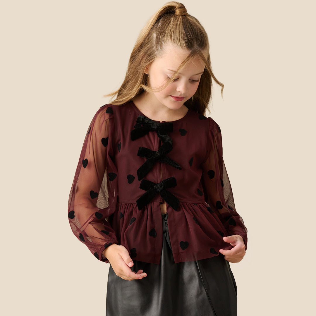 A young girl with blonde hair in a high ponytail is wearing a dark burgundy sheer long-sleeve blouse with black heart and bow details, paired with a black skirt, against a solid beige background.