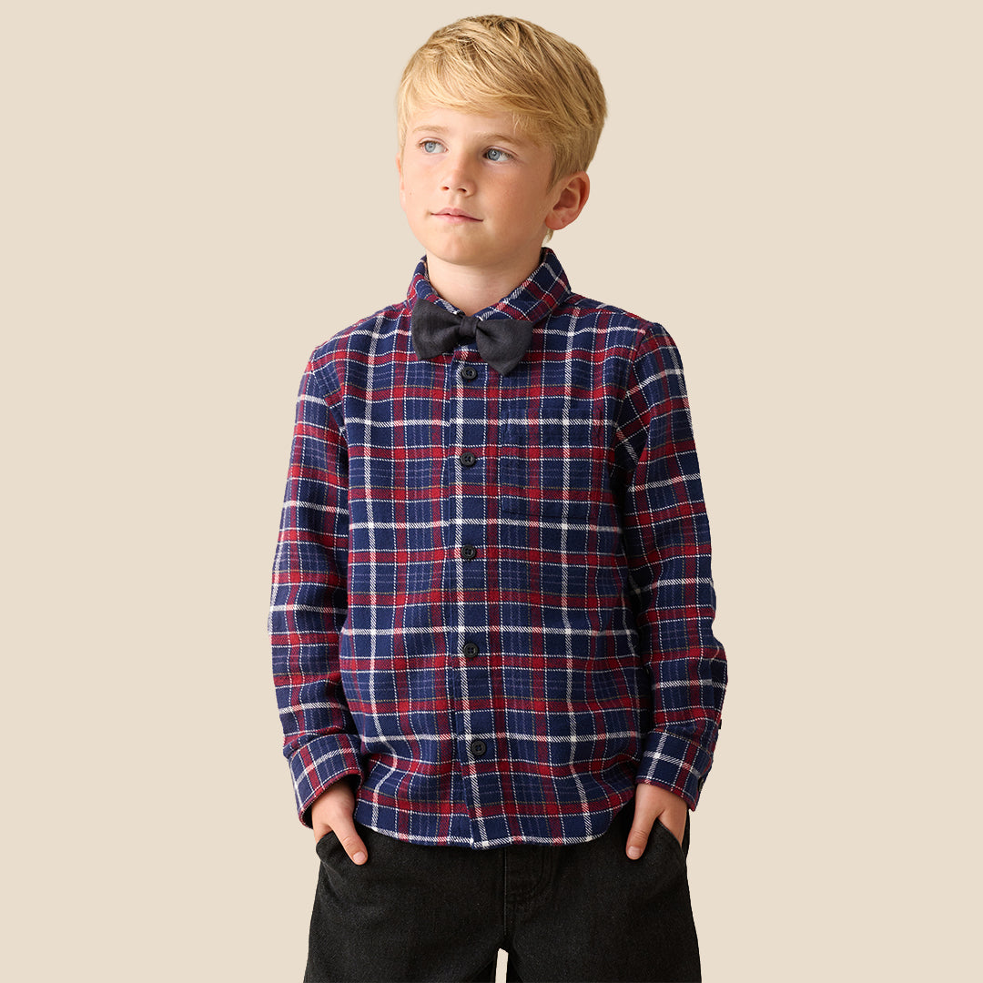 A young boy with blond hair wearing a red, navy, and white plaid long-sleeve button-up shirt with a black bow tie, paired with black pants, standing against a solid beige background.