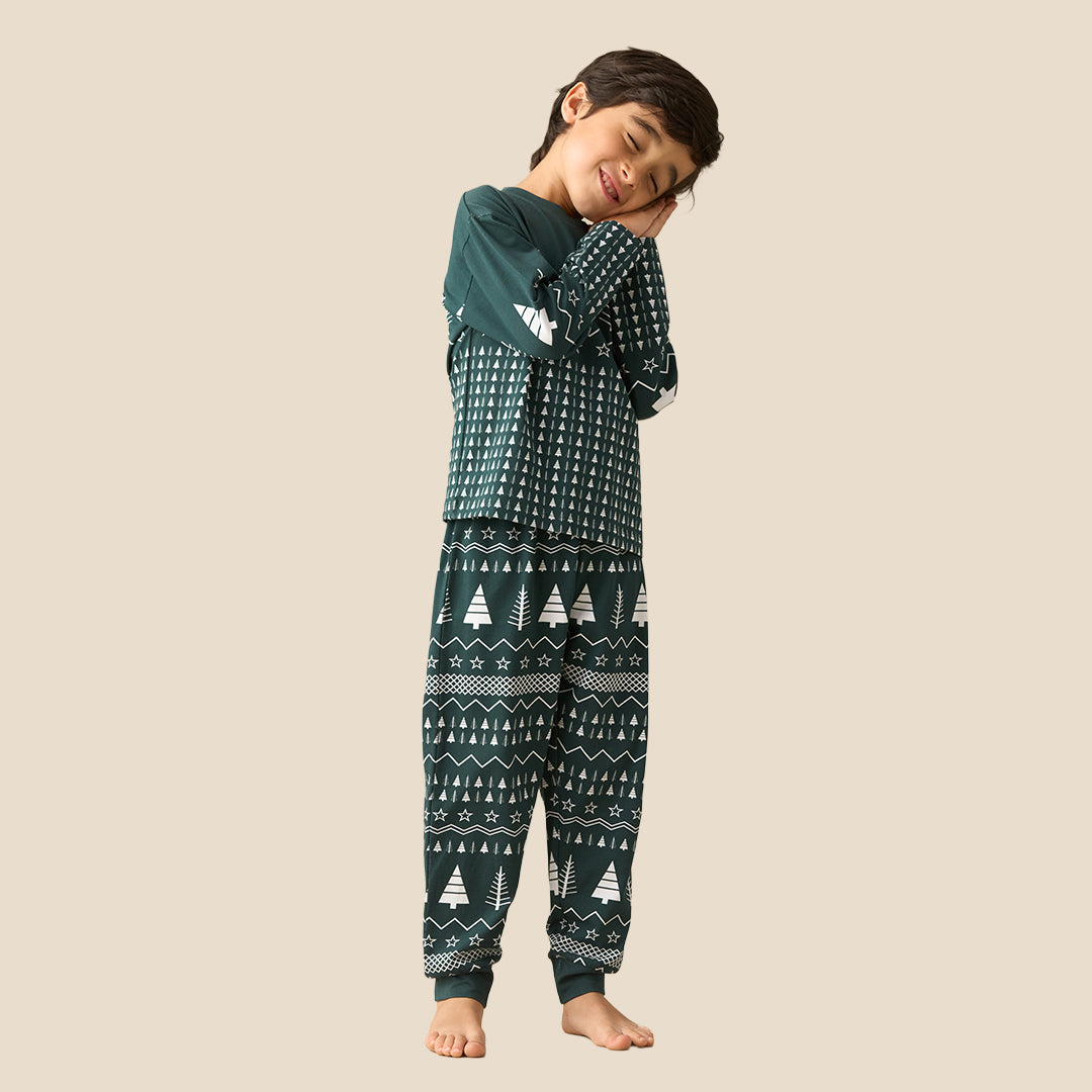 A young boy stands barefoot against a solid beige background, wearing a dark green long-sleeve pajama set with a white Nordic tree pattern.