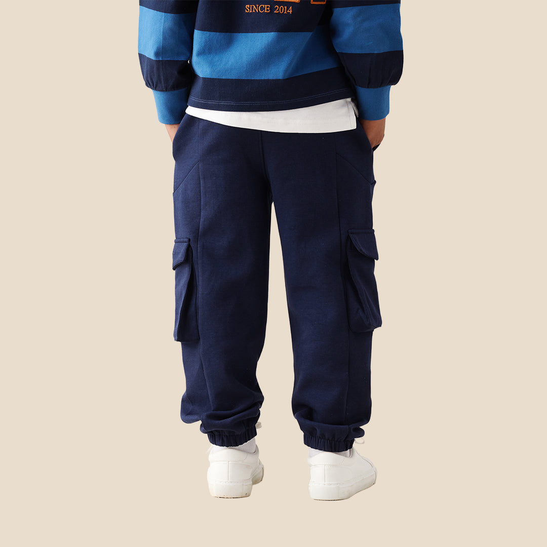 A child wearing navy blue cargo pants with side flap pockets, a blue and navy striped long-sleeve shirt, and white sneakers, standing against a solid beige background.