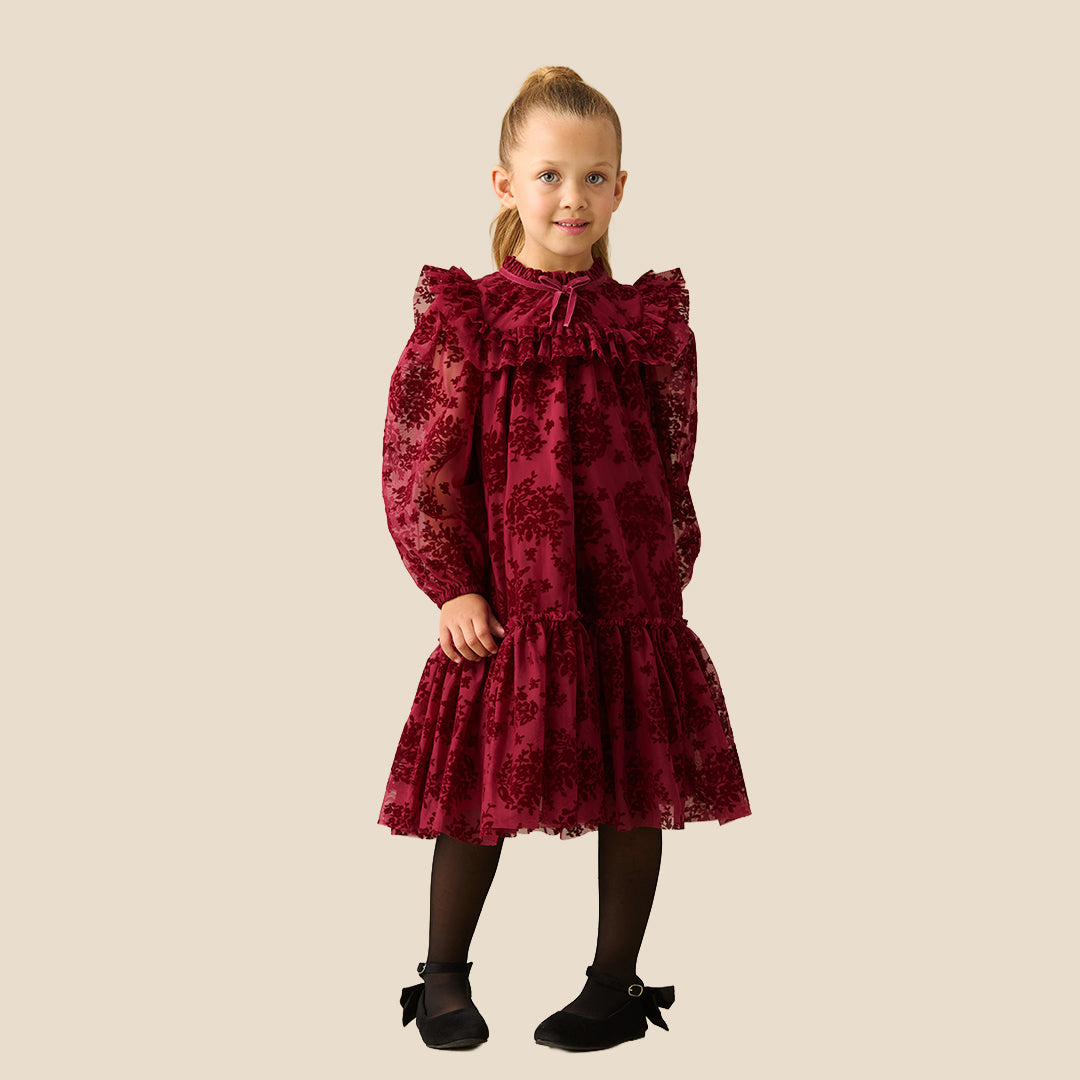 A young girl with her hair in a ponytail models a long-sleeved burgundy dress with a floral lace overlay, ruffled collar, and tiered skirt, set against a solid beige background.