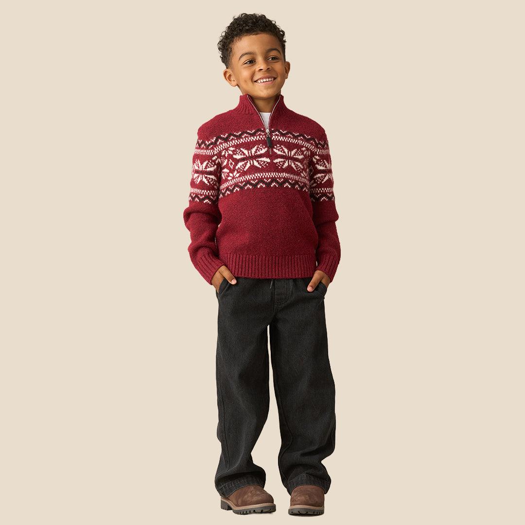 A young boy smiling and wearing a burgundy knit quarter-zip sweater with a white and dark red Fair Isle snowflake pattern across the chest and shoulders, set against a solid beige background.