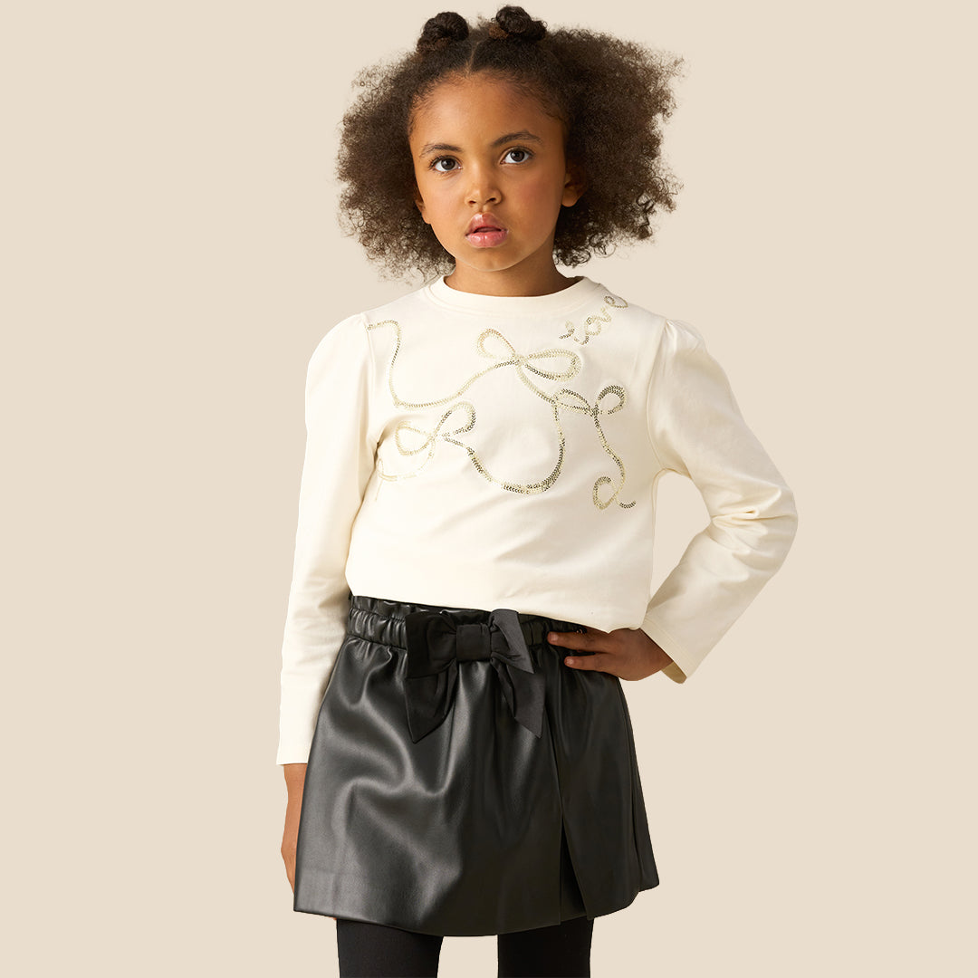 A child wearing a black faux-leather wrap mini skirt with a large black fabric bow at the waist, styled with a cream long-sleeve top and black tights against a solid beige background.