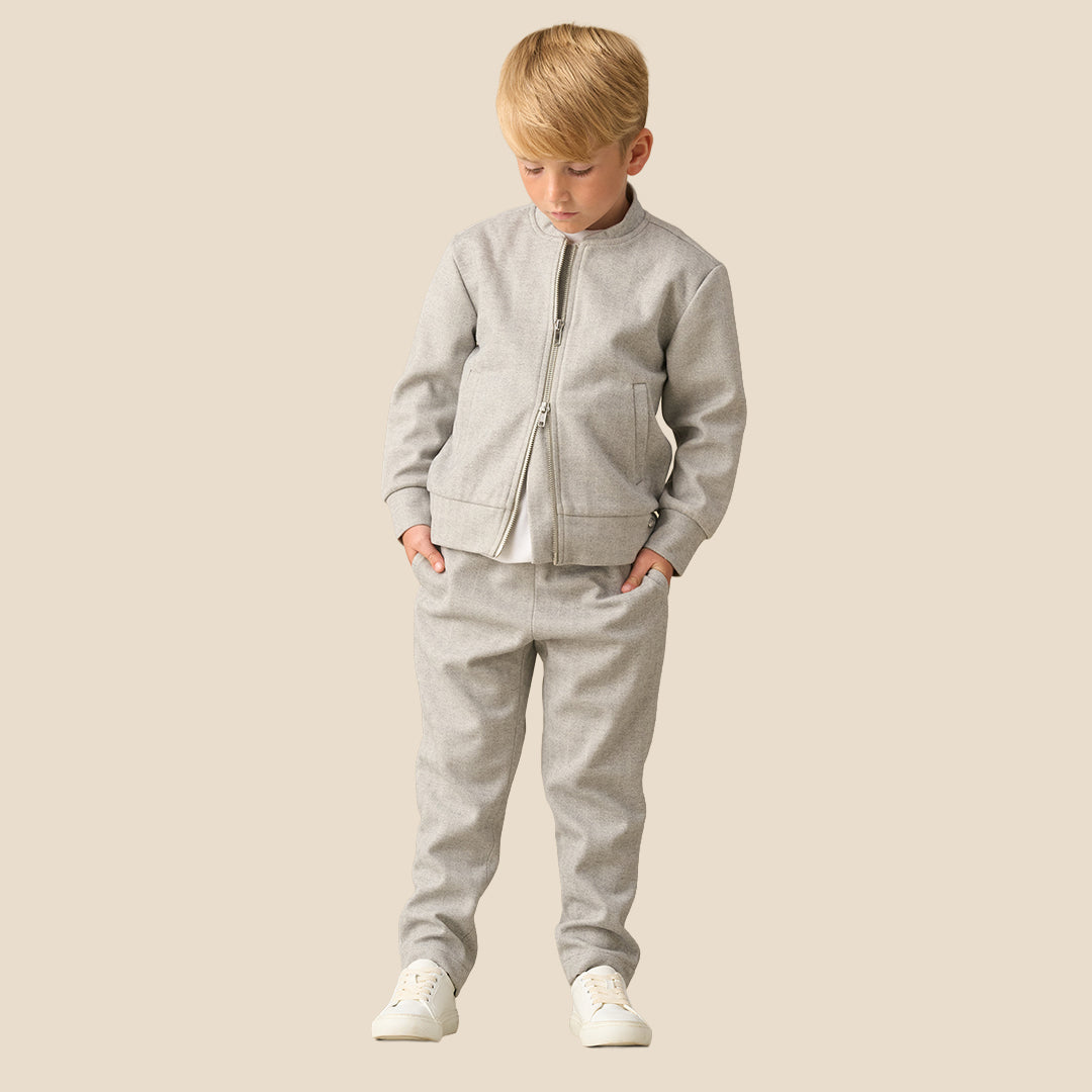 A young boy with blond hair wearing a light gray zip-up fleece jacket and matching gray sweatpants, standing against a solid beige background.