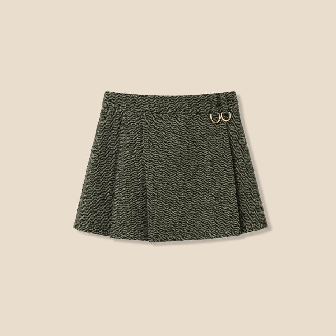 A model wearing a light pink sweatshirt with brown cursive text and a dark olive green pleated tweed mini skirt, standing against a solid beige background.