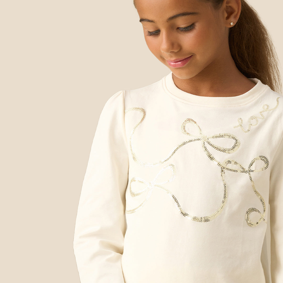 A young girl with her hair in a ponytail is wearing a cream-colored long-sleeve top with metallic gold sequin embroidery and shiny gold sequin pants, set against a solid beige background.