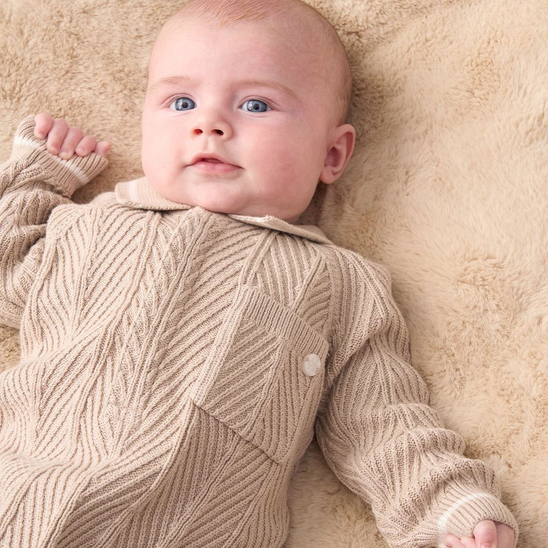 A baby wearing a beige and white textured knit one-piece jumpsuit with white shoes, standing against a solid beige background.