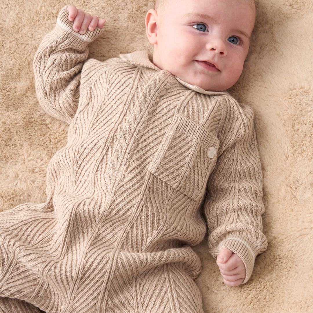 A baby wearing a beige and white textured knit one-piece jumpsuit with white shoes, standing against a solid beige background.