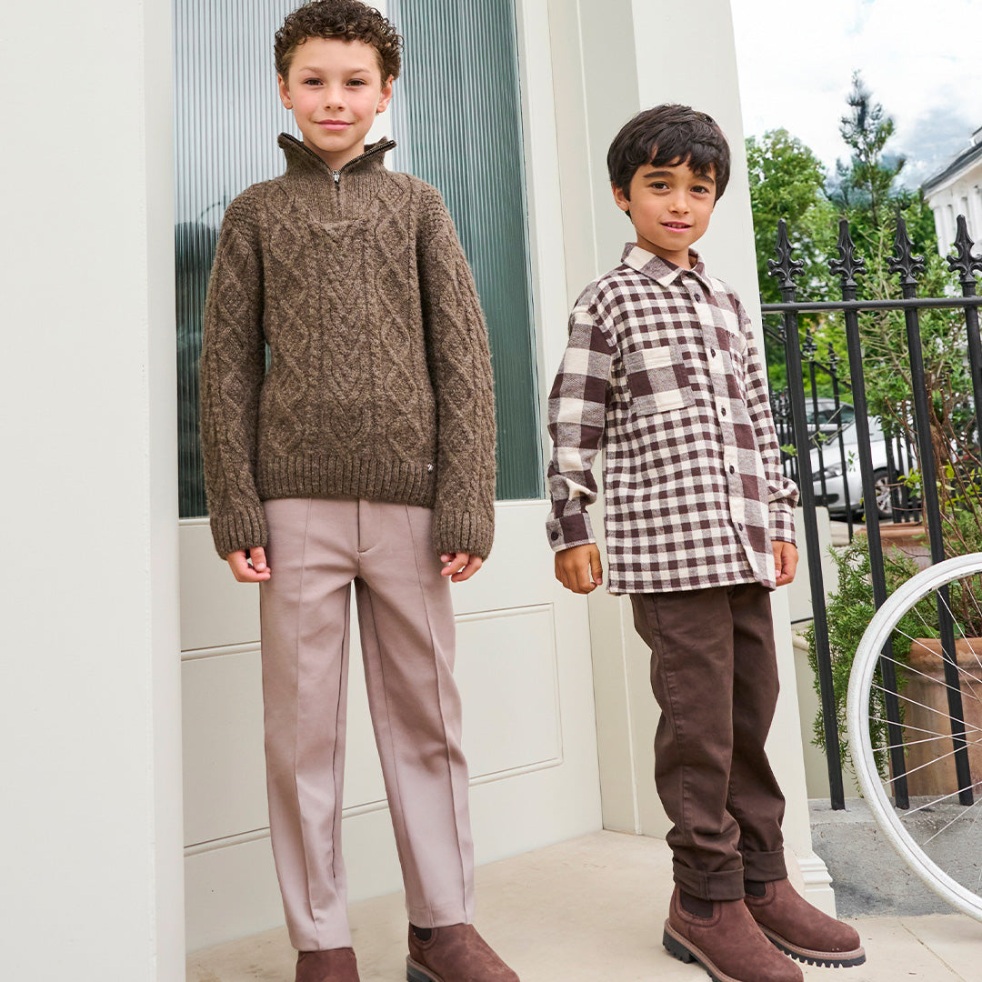A child wearing a brown cable-knit mock-neck sweater and light pink trousers, standing indoors near a white wall and window. Another child in a brown-and-white plaid shirt is partially visible in the foreground.