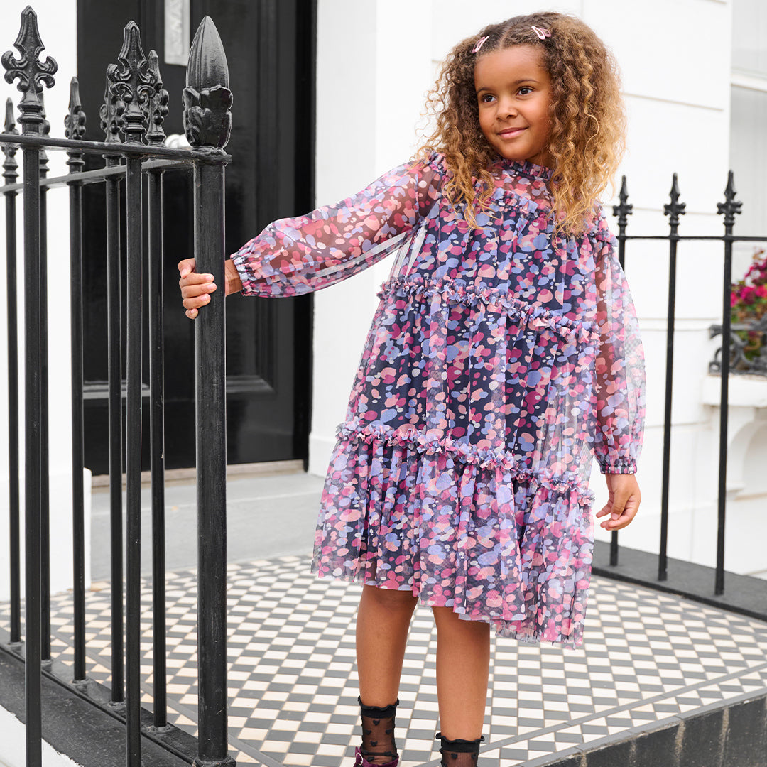 A young girl with curly hair stands on a checkered porch wearing a long-sleeved tiered dress with a pink, purple, and blue floral pattern. She is posing behind a black wrought-iron fence.
