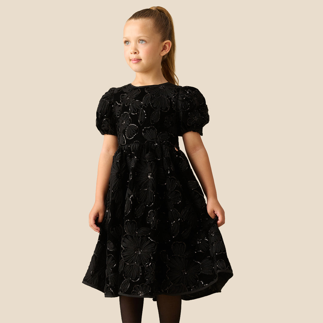 A young girl with her hair in a ponytail models a black short-sleeve dress with textured floral applique, standing against a solid beige background.