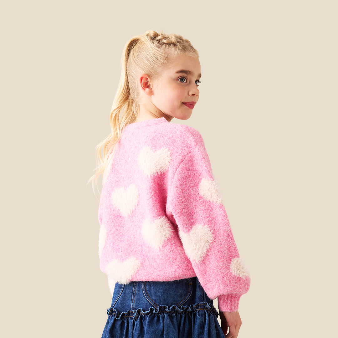 A young girl with blonde hair styled in buns wears a pink fuzzy knit sweater with fluffy white heart details, posing with her hands forming a heart shape in front of her chest. She is wearing a dark blue denim skirt.