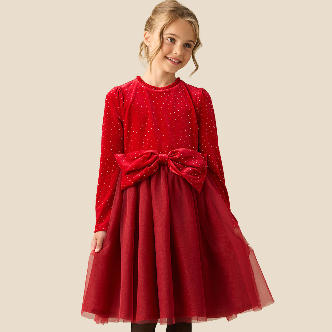 A young girl wearing a festive red dress with long sleeves, tiny gold polka dots, a matching red bow at the waist, and a tulle skirt, set against a solid beige background.