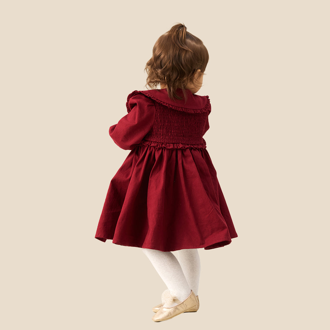 A toddler girl wearing a burgundy corduroy dress with a large white ruffled collar decorated with bows, white tights, and beige ballet flats, standing against a solid beige background.