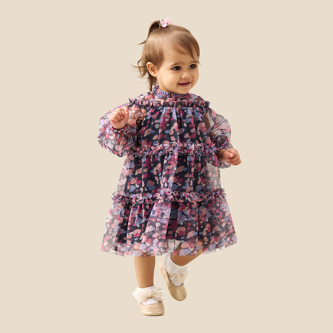 A toddler girl wearing a tiered long-sleeve dress with a dark navy base and a colorful floral pattern in pink, purple, and blue, standing against a solid beige background.