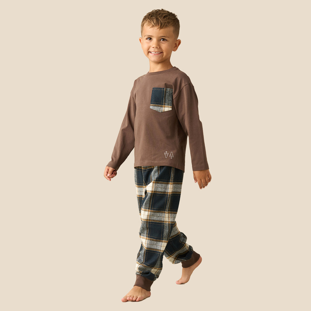 A young boy modeling a two-piece pajama set consisting of a brown long-sleeve shirt with a plaid chest pocket and matching plaid pants, set against a solid beige background.