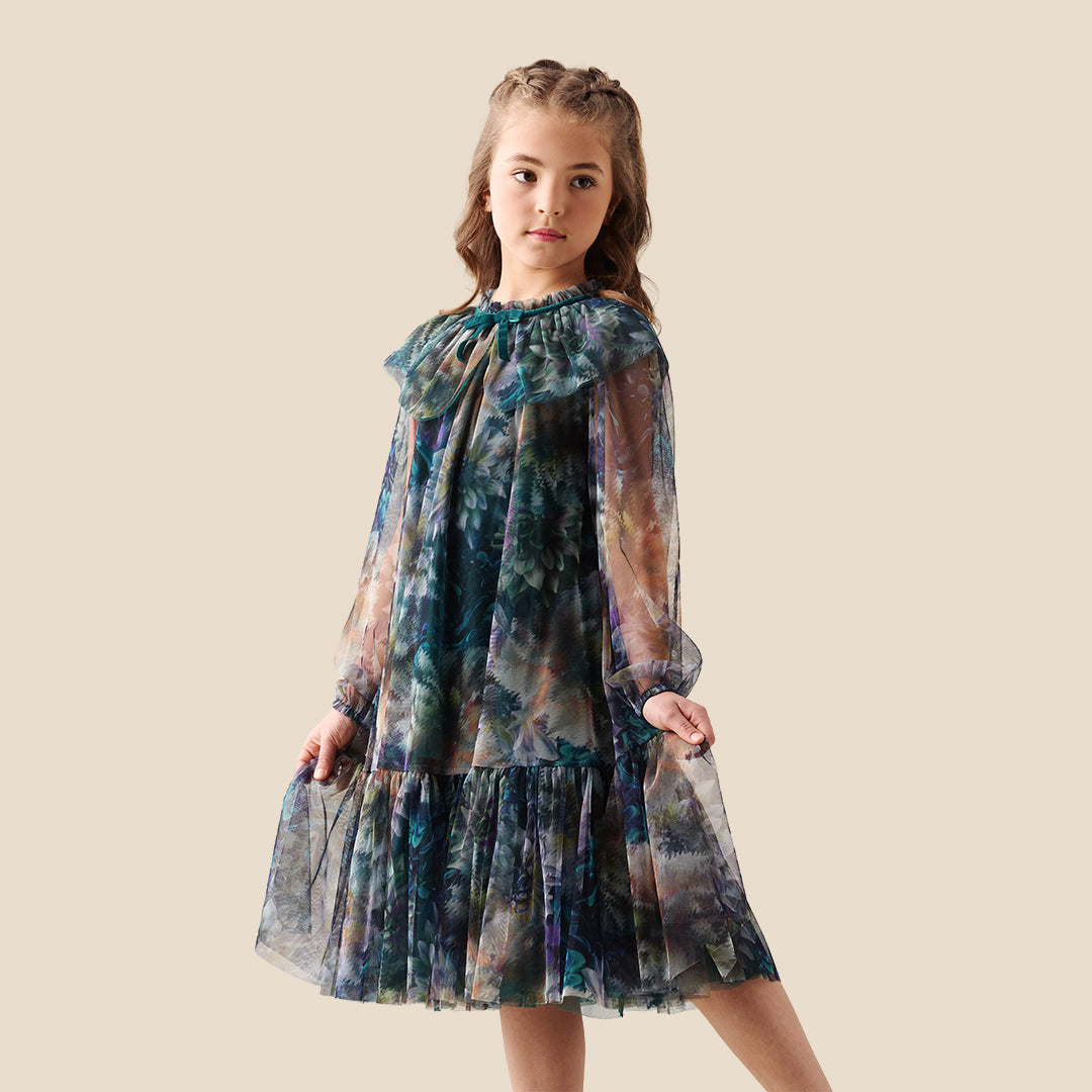 A young girl modeling a sheer, tiered tulle dress with a large ruffled collar and a blue, green, and purple floral watercolor pattern, set against a solid beige background.