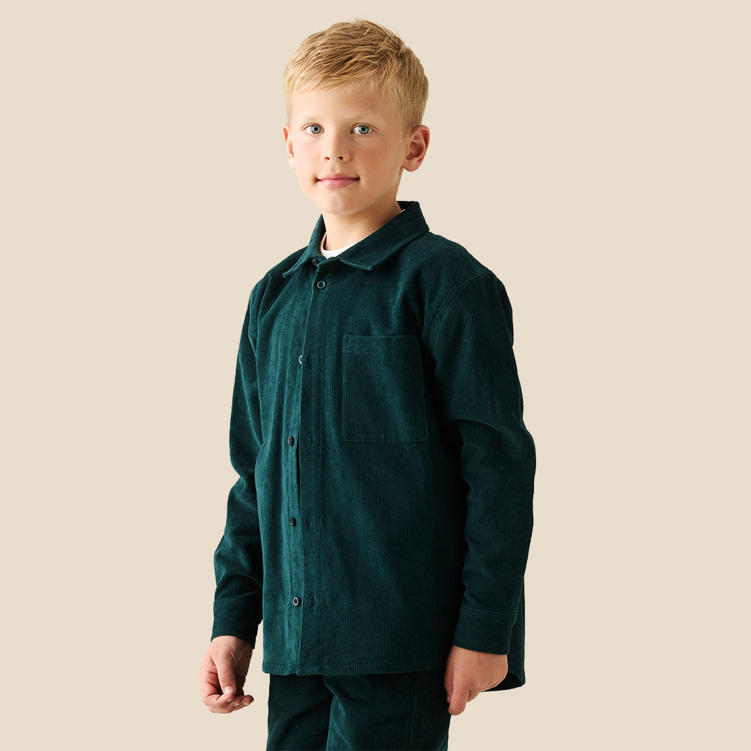A young boy with blonde hair wearing a dark forest green corduroy button-up overshirt over a white t-shirt and matching green pants, standing against a solid beige background.