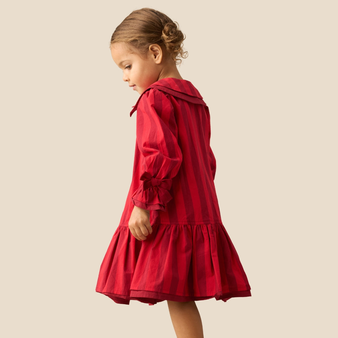 A young girl with her hair in a bun is wearing a red and dark red vertically striped long-sleeve dress, shown from a side-back angle against a solid beige background.