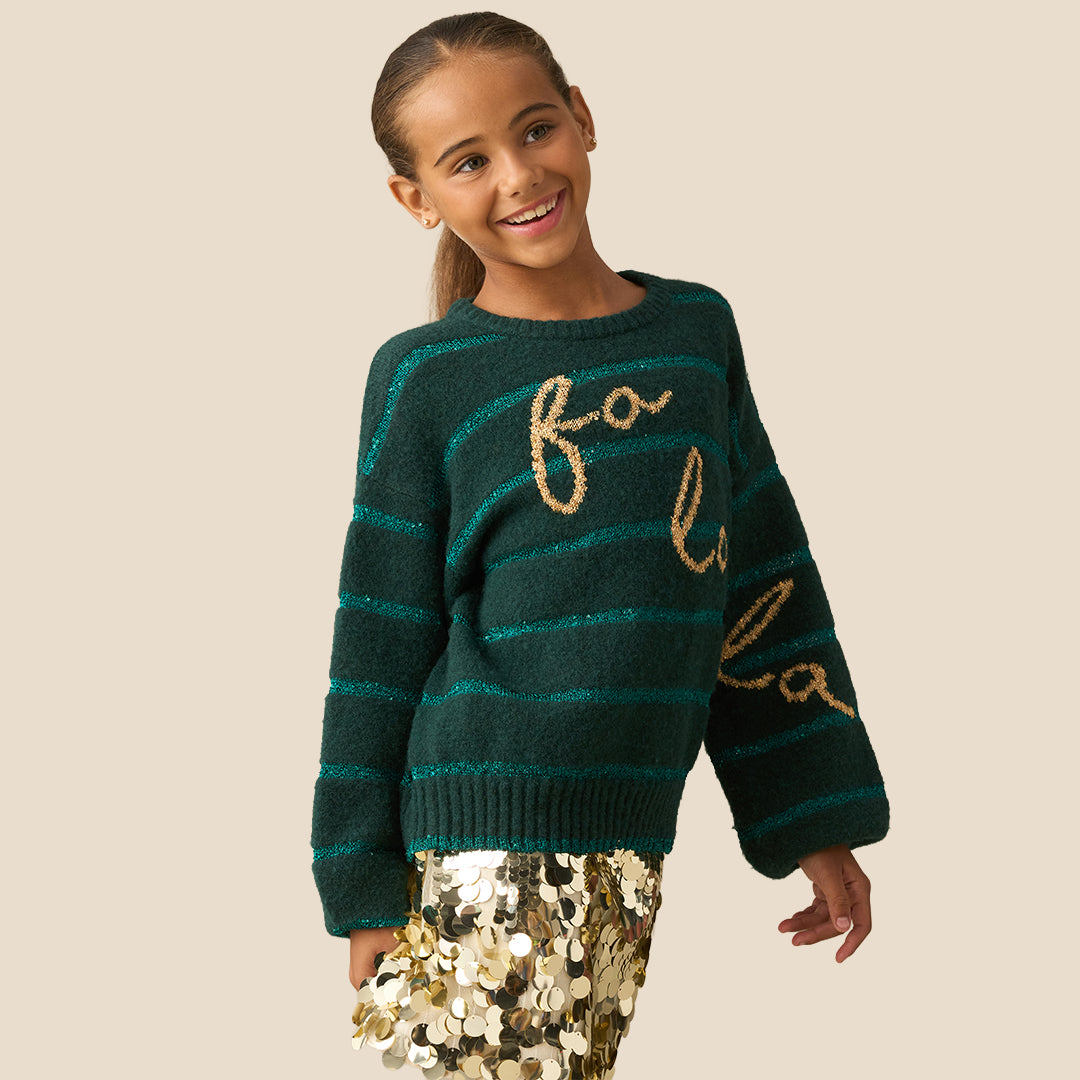 A young girl smiling while wearing a dark green knit sweater with teal horizontal stripes and gold glittery 'fo'lo' lettering on the front, paired with a gold sequined skirt, against a solid beige background.