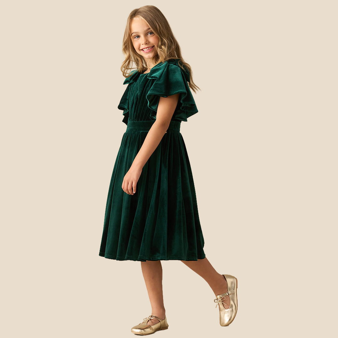 A young girl wearing a dark green velvet dress with ruffled short sleeves, a matching waist belt, and a pleated skirt, standing against a solid beige background.