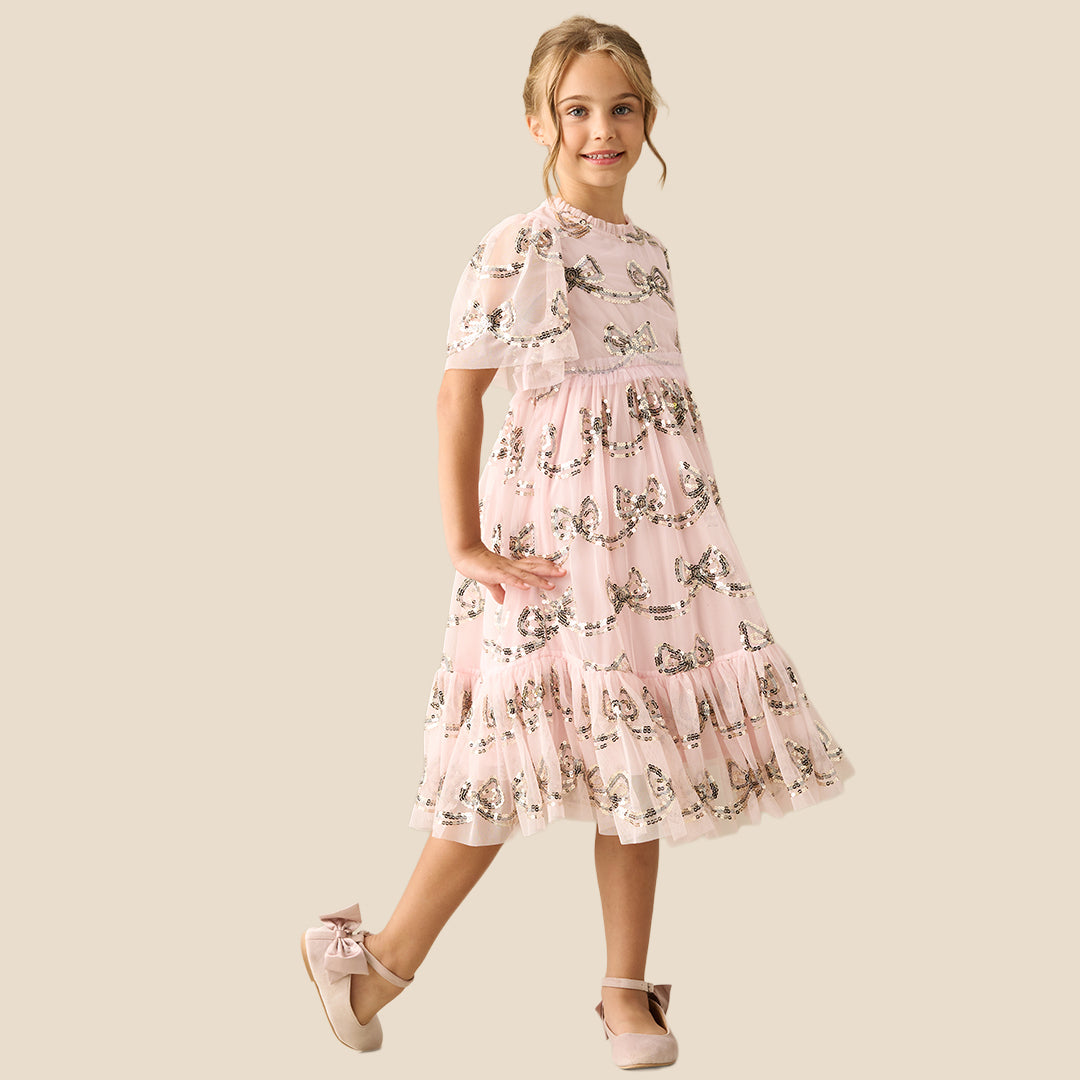 A young girl smiling and wearing a light pink tiered tulle dress with a delicate black and white floral print, set against a solid beige background.