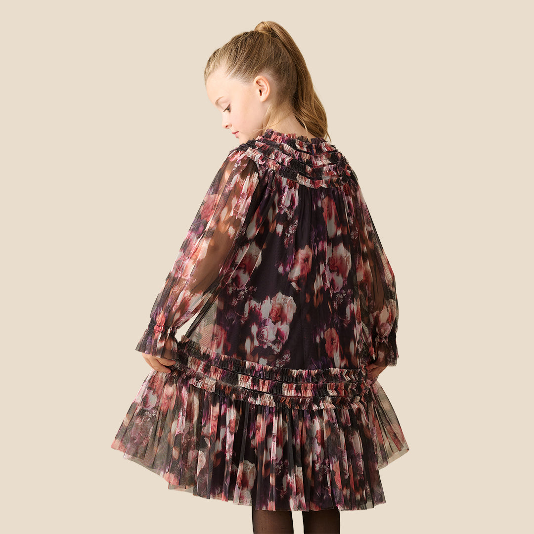 A young girl with her hair in a ponytail is wearing a long-sleeved, tiered tulle dress with a pink, red, and white floral pattern on a dark background, set against a solid beige studio backdrop.