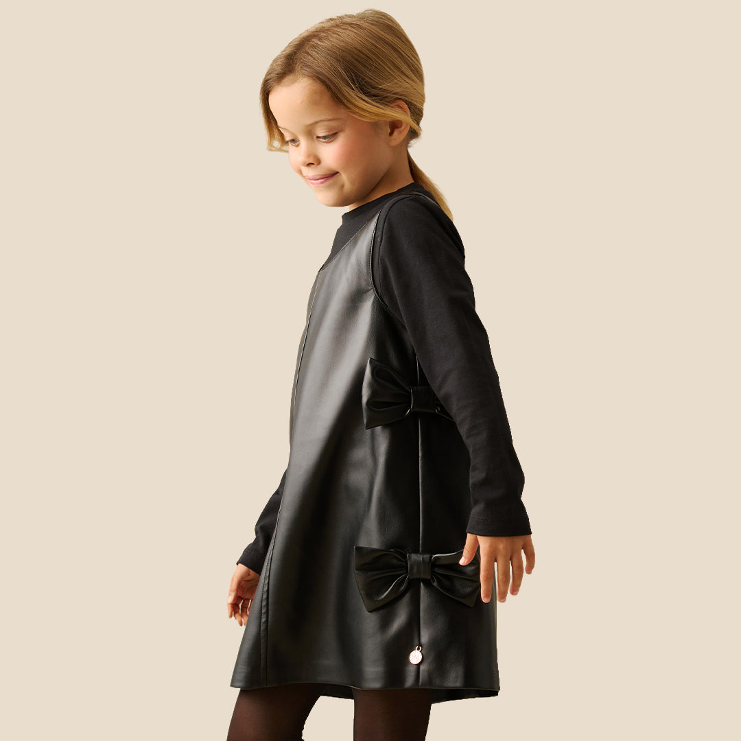 A young girl with blonde hair tied back is wearing a black faux-leather dress with long sleeves, a mock neck, and decorative bows on the sides, paired with black tights, against a solid beige background.