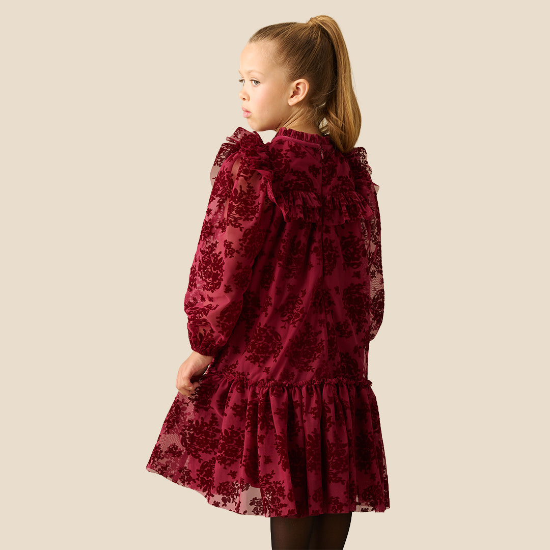 A young girl with her hair in a ponytail models a long-sleeved burgundy dress with a floral lace overlay, ruffled collar, and tiered skirt, set against a solid beige background.