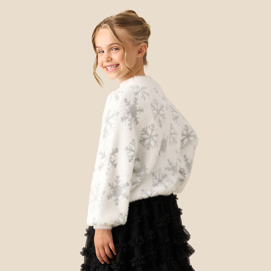 A young girl with blonde hair in a bun wearing a fuzzy white long-sleeve sweater with a gray snowflake pattern, paired with a black ruffled skirt, set against a solid beige background.