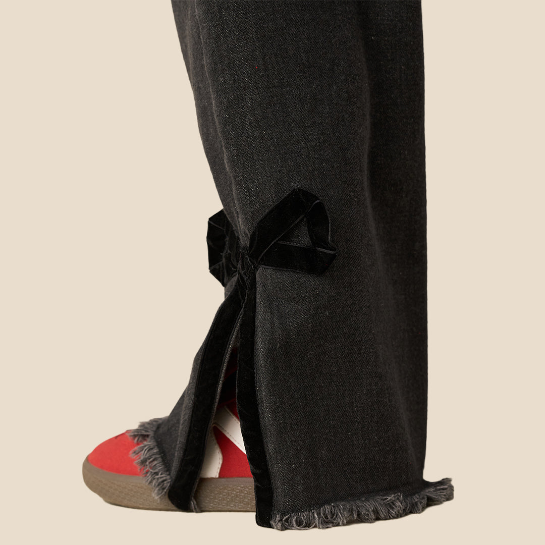 A person wearing black wide-leg denim jeans with side tie details and raw frayed hems, styled with a red bow-tie sweatshirt and red sneakers, against a solid beige background.