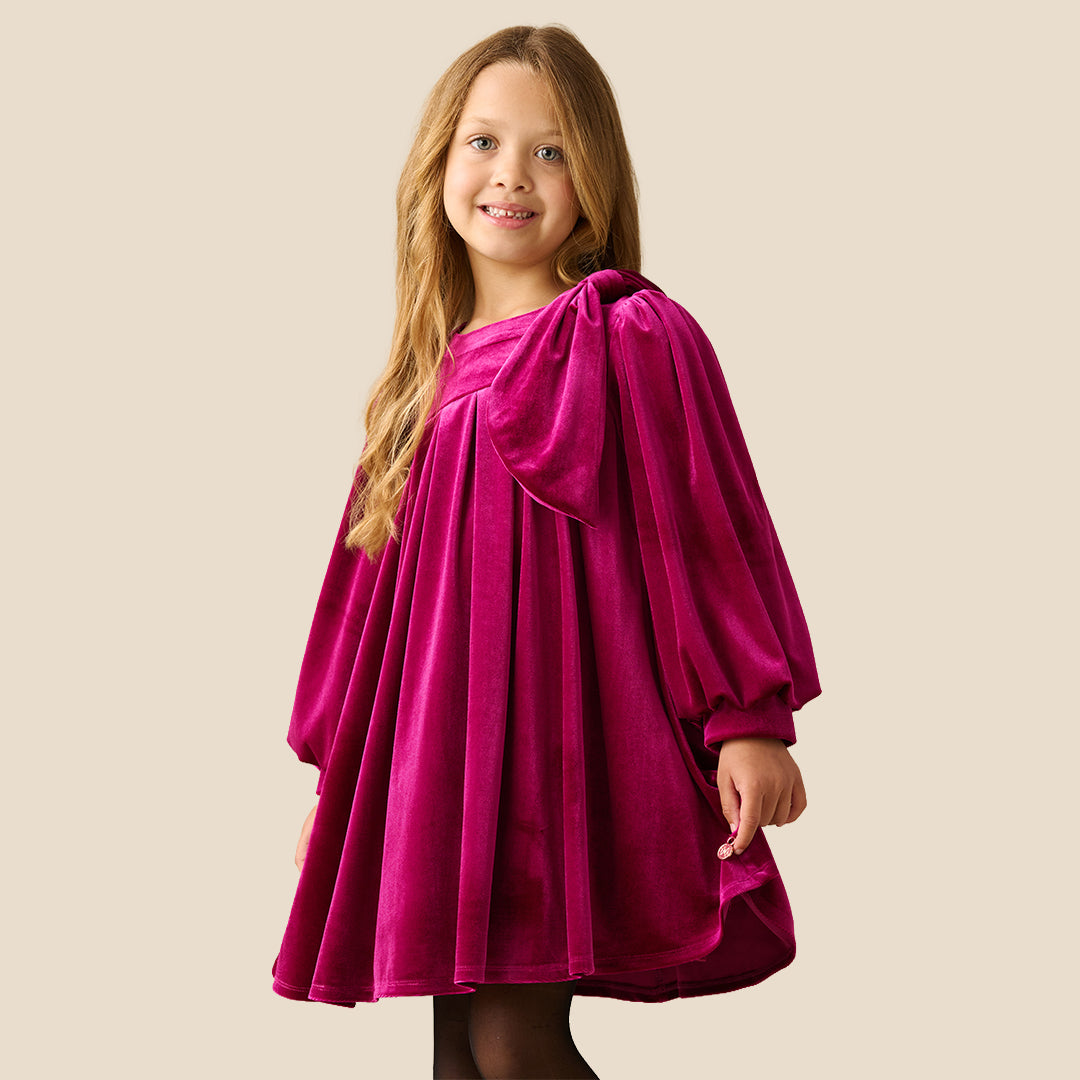 A young girl with blonde hair wearing a long-sleeved magenta velvet dress with a gathered bow detail on the chest, standing against a solid beige background.