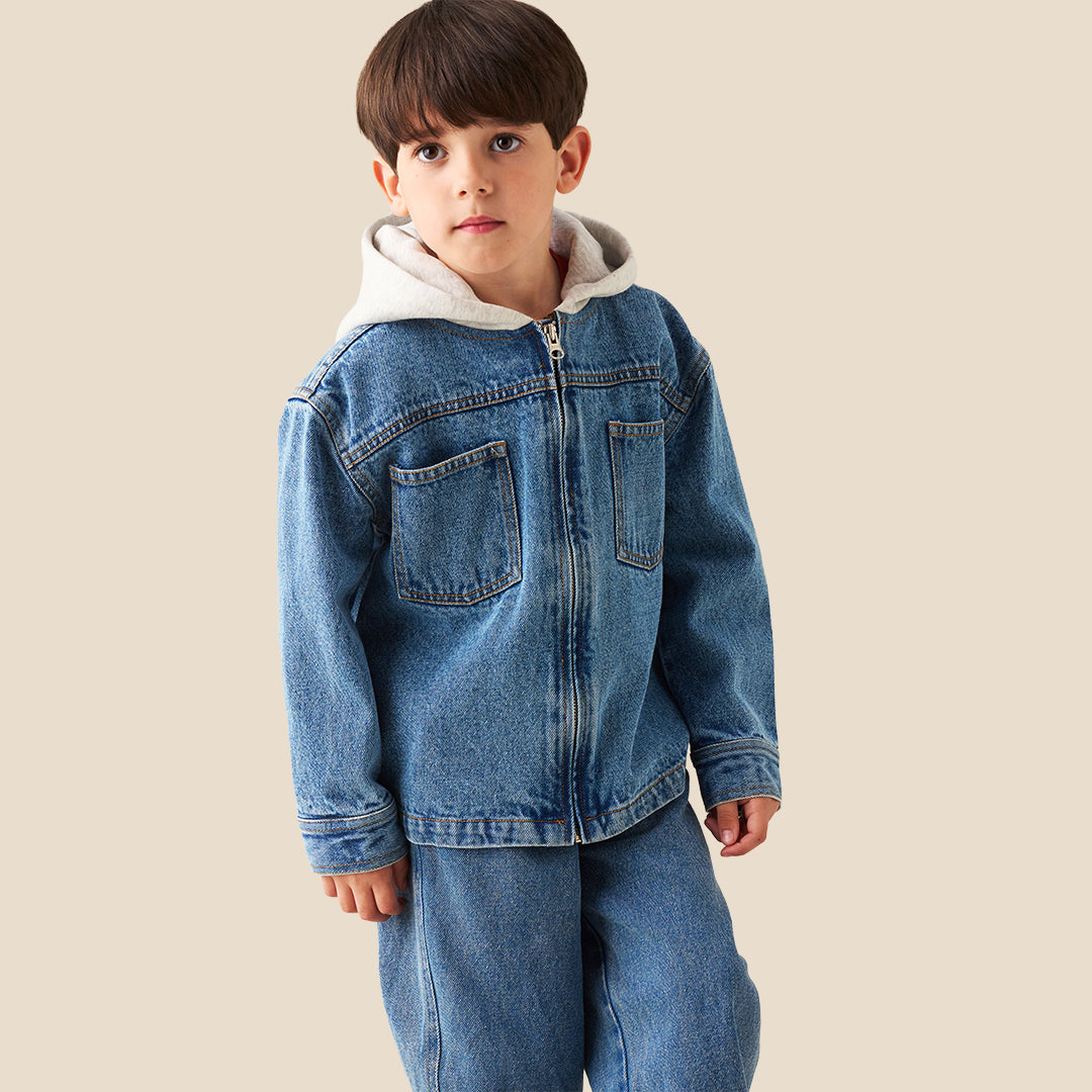 A young boy wearing a light blue denim zip-up jacket with a white hood lining and matching blue jeans, standing against a solid beige background.