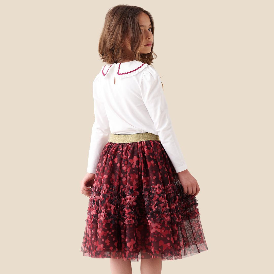 A child wearing a white long-sleeve top and a tiered tulle skirt in dark red and black floral print with a gold waistband, shown against a plain beige background.