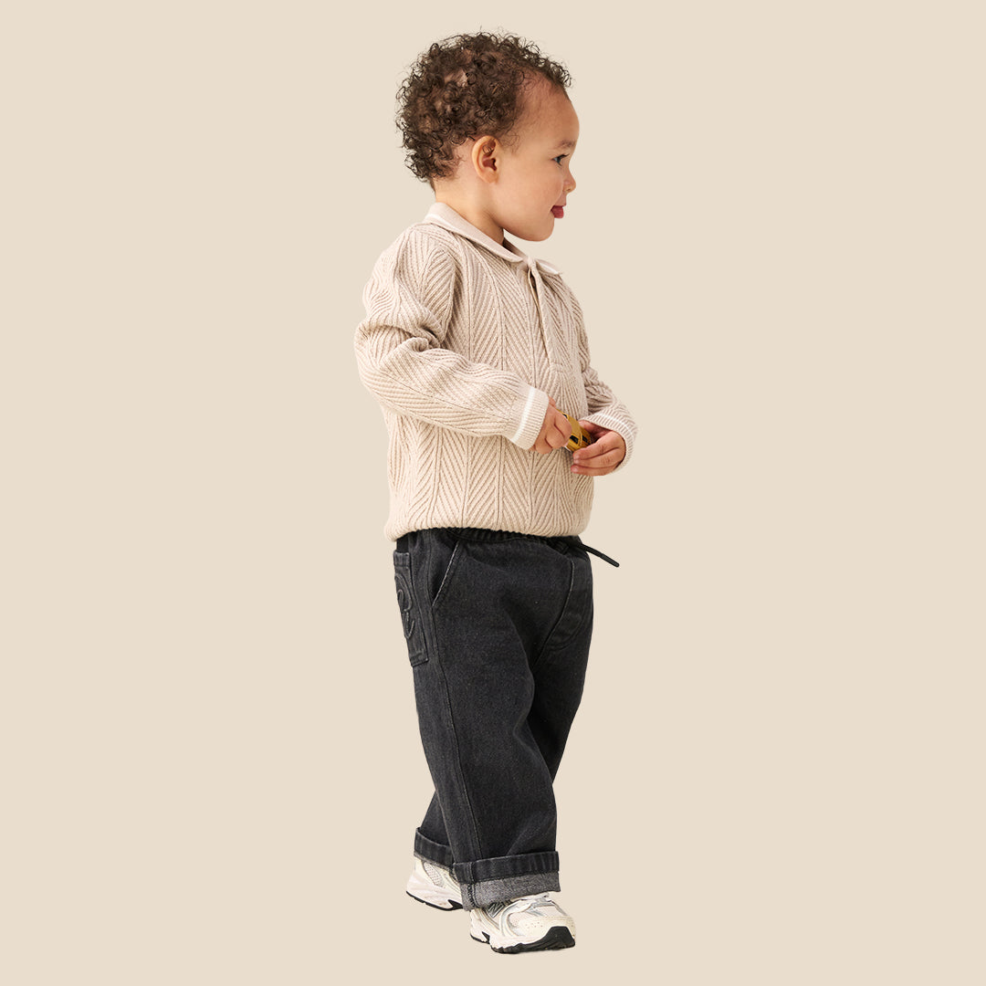 A toddler with curly brown hair wearing a cream-colored cable-knit sweater with a white Peter Pan collar, standing against a solid beige background.