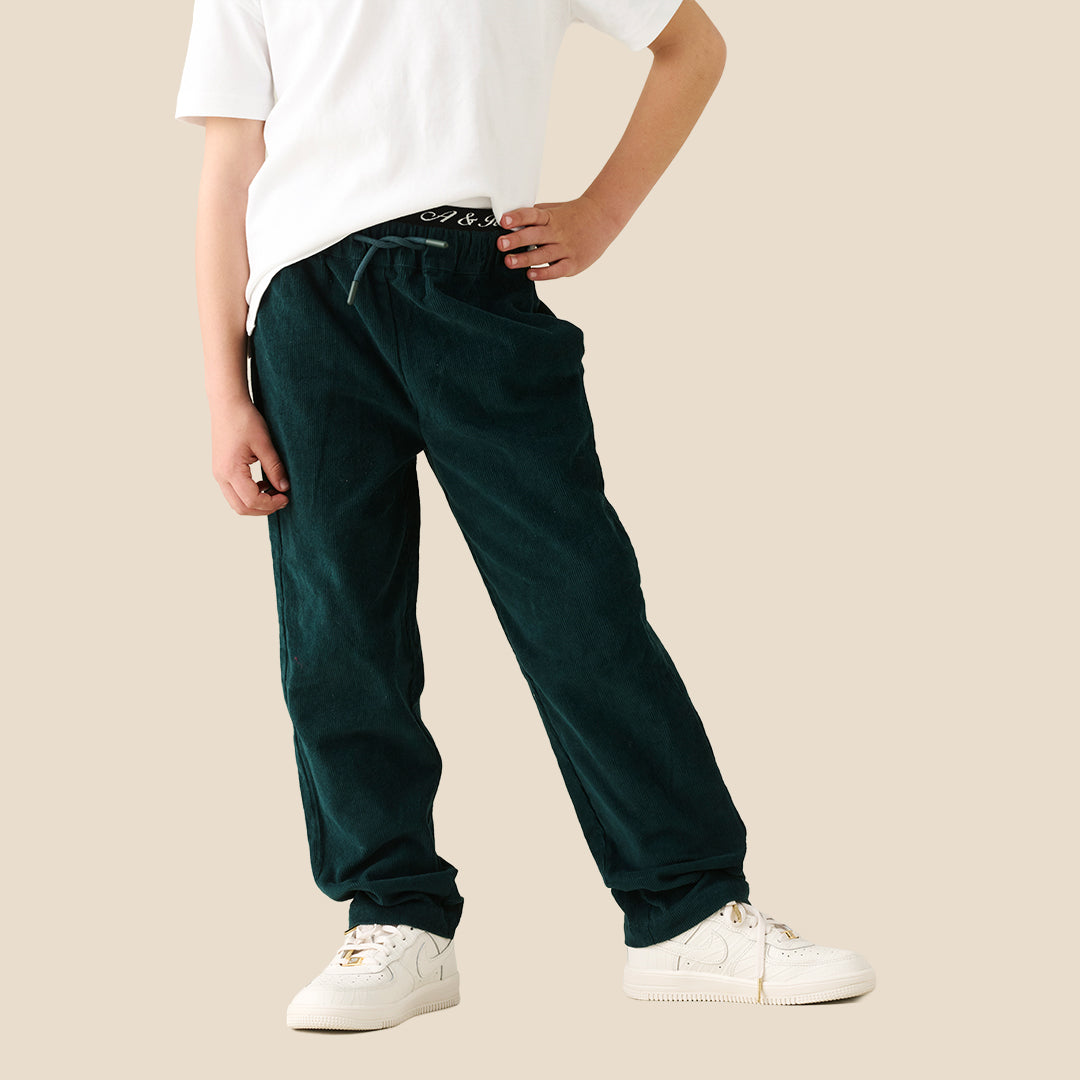 A young boy modeling a white crewneck t-shirt and dark green corduroy pants, standing against a solid beige background.