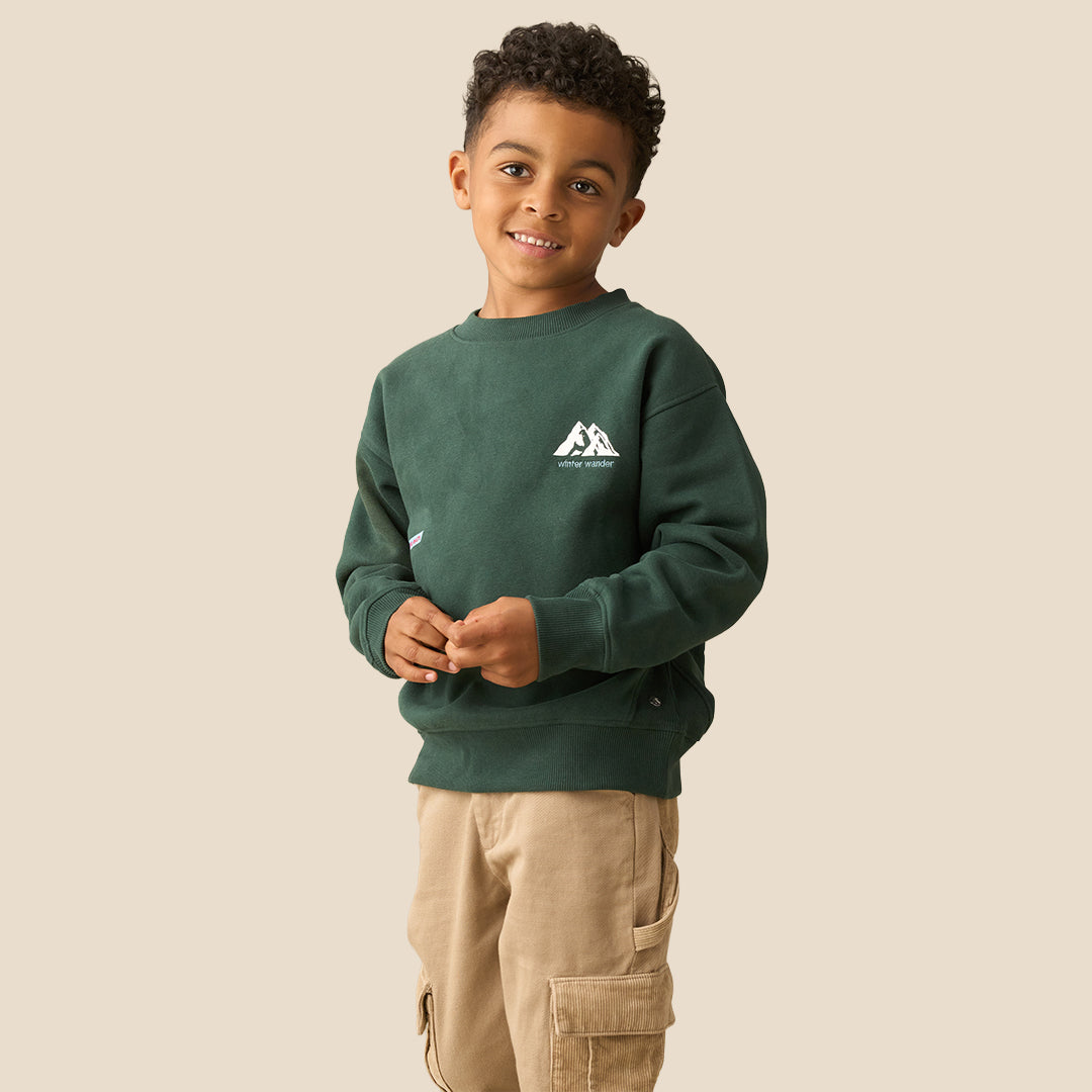 A young boy with curly dark hair wearing a dark green crewneck sweatshirt with a small white graphic on the left chest and khaki pants, posing against a solid light beige background.